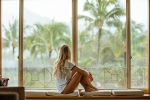 девушка сидит у окна в дождливый день, пальмы, фото со спины 
