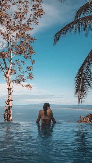 Девушка в бассейне отеля с видом на Тихий океан, деревья, пальма, голубое небо 
