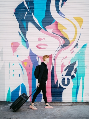 человек с чемоданом идет вдоль стены с ярким граффити в виде девушки 