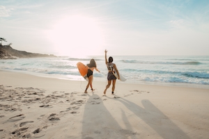 две девушки с серфбордами идут по пляжу в сторону моря 