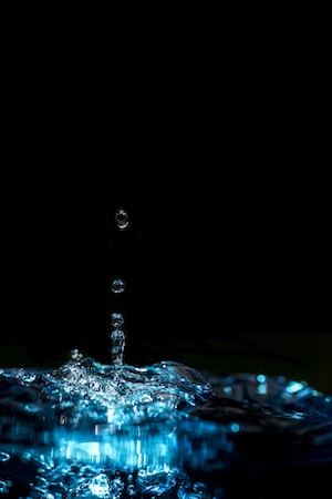 капля воды отскакивает от водной поверхности, крупный план, макро-фотография, вода светится голубым светом на черном фоне 