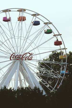 Съемка ярмарочного колеса на ярмарочной площади в Салониках, Греция. На ярмарочном колесе изображен логотип Coca-Cola, и несколько человек весело проводят время, катаясь на ярмарочном колесе.