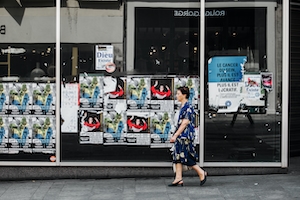 Пожилая женщина идет по улице мимо постеров, плакатов на стене 