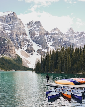 Горное озеро, отражение неба и гор в воде, лес у озера и гор, человек стоит на пирсе около небольших лодок 