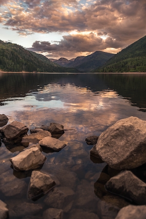 Горное озеро, отражение неба и гор в воде, лесу у озера и гор, большие камни на озере во время заката 