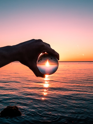 закат над водой, красочное солнце и небо, солнечная дорожка по воде, стеклянный шар в руке человека 