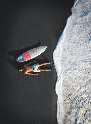 фото морского побережья с темным песчаным пляжем с высоты, парень лежит на песке у доски для серфинга 
