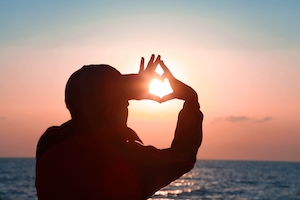 Силуэт человека, показывающего сердце руками, на фоне заката, красочное солнце и небо