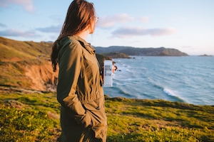 девушка на зеленом скалистом побережье держит в руках белую коробку с водой 