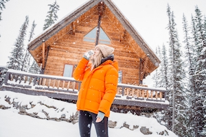 девушка в оранжевой куртке пьет на фоне деревянного дома зимой 
