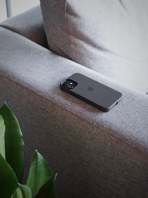 iPhone 12 Mini с чехлом Apple на сером льняном диване лицом вниз с растением на переднем плане.