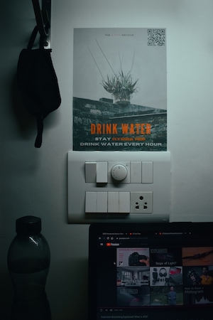 Открытый ноутбук с запущенным YouTube, рядом с ним бутылка воды и висящая маска. На стене висит плакат, советующий пить воду.