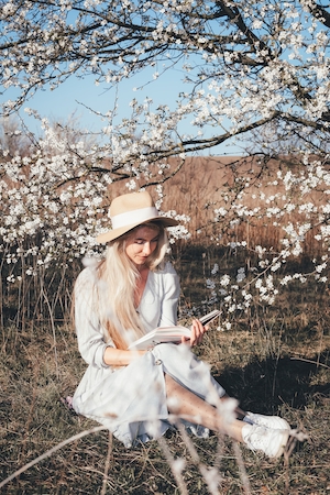 Блондинка с длинными волосами, молодая женщина читает книгу. Девушка, портрет весной в парке, вокруг цветут деревья и распускаются цветы сакуры пастельных тонов. Цветущие ветки дерева сакура. 