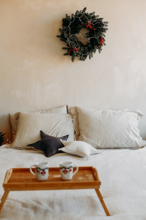 Новогодний декор, украшенный еловый венок на стене, заправленная постель, новогодние кружки на маленьком столике, завтрак в постель 