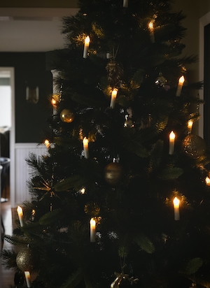 Фотография рождественской елки крупным планом, украшенной гирляндами в форме свечей и разными золотыми украшениями. На заднем плане - темно-зеленые стены с золотистыми настенными бра.
